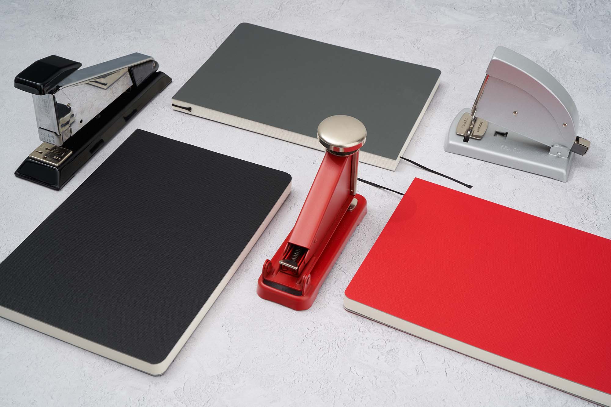 the paper mind Mitsubishi Bank paper notebooks in red gray and black laying flat on a gray background with black red and gray staplers, notebooks for fountain pens