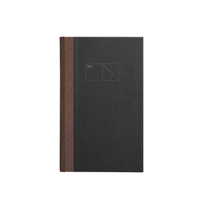A minimalist hardcover notebook or journal standing upright on a light gray background. The cover is a very dark charcoal gray with a thin vertical brown spine band and a small geometric line drawing printed near the top center. Papierniczeni Copper 2027 Calendar – Graphite