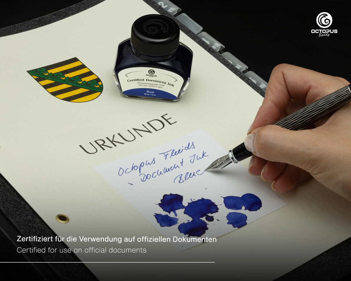 A hand holds a silver fountain pen over a white document, writing blue ink. A small bottle of Octopus Ink document ink sits nearby on a dark surface, with blue ink swatches and the word 'URKUNDE' visible on the page. Octopus Fluids Fountain Pen Document Ink - 30 ml Bottle - Dark Blue -  Made in Germany