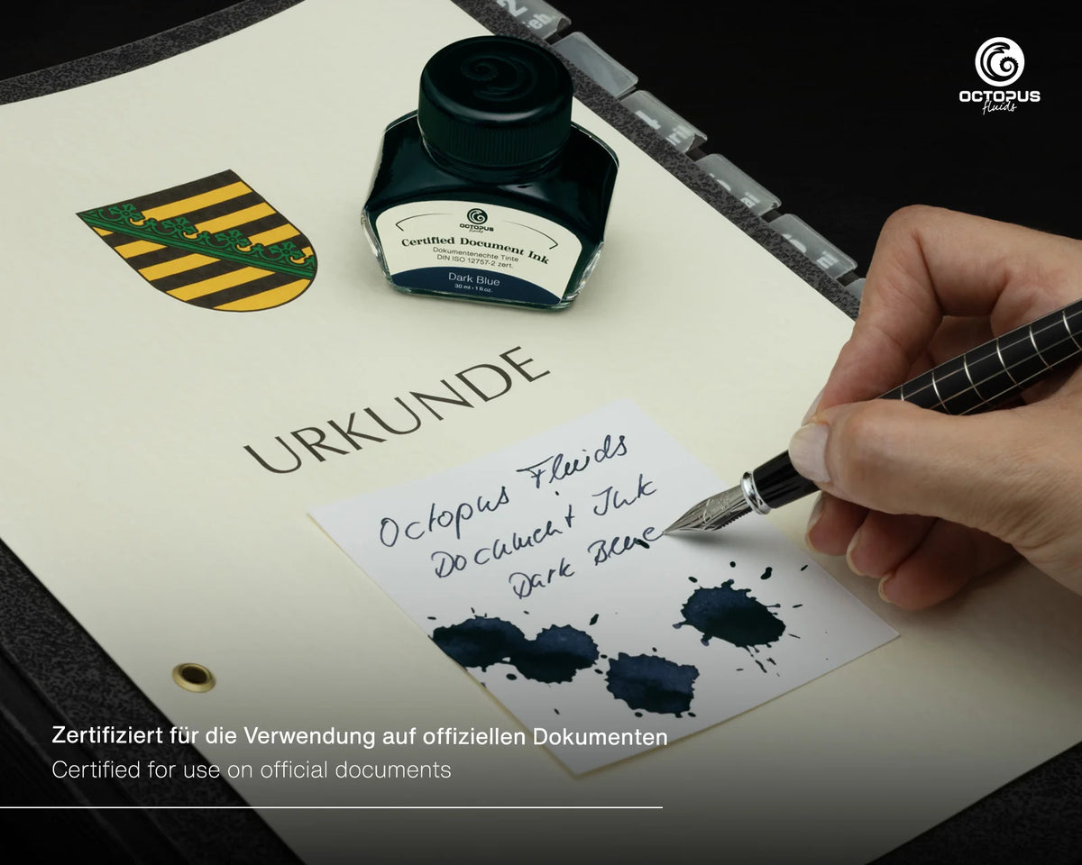 Octopus Fluids Document Ink in Dark Blue, shown in its glass bottle next to a hand writing with a fountain pen on an off-white document. The document also features a printed crest and the word 'URKUNDE'. Octopus Fluids Fountain Pen Document Ink - 30 ml Bottle - Dark Blue -  Made in Germany