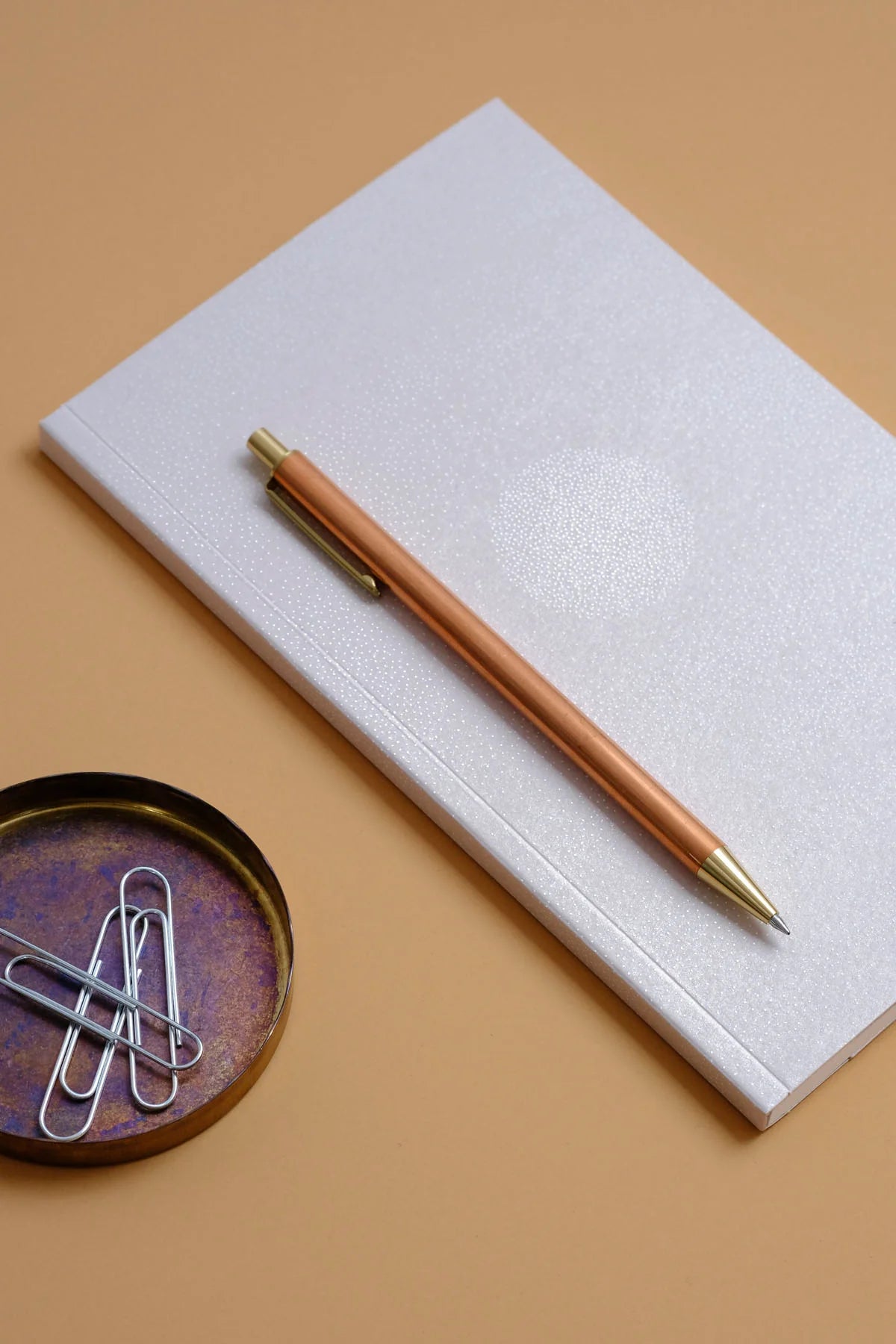 A rose gold metallic ballpoint pen with gold accents rests diagonally on a textured white notebook, set against a warm beige background with a small decorative dish of paperclips in the lower-left corner. Papierniczeni Luna Notebook - Full Moon