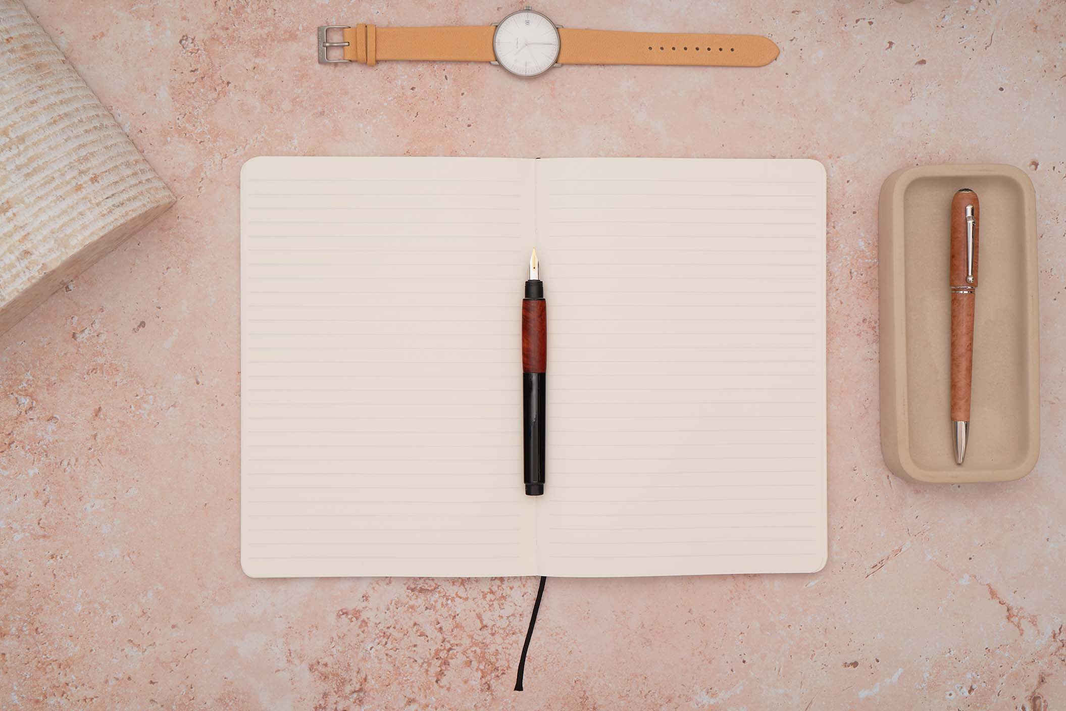 the paper mind Mitsubishi Bank paper notebook lined ruled open on a pink stone background with a Lamy accent fountain pen in the middle a junghans watch on top and a dunhill sidecar briarwood ballpoint in a concrete tray