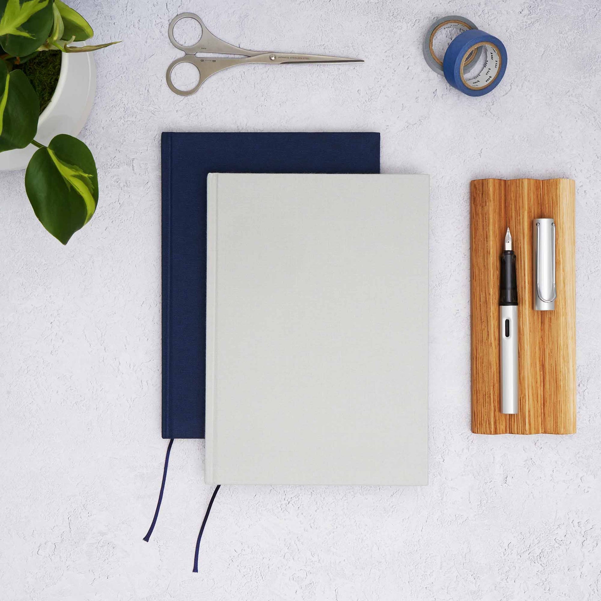 The Paper Mind Cosmo Air Light Hardcover Notebooks in grey and blue with an oak wood pen tray, Lamy AL Star fountain Pen, MT washi tape, Before Breakfast London scissors and plant. Fountain pen friendly notebooks from the paper mind