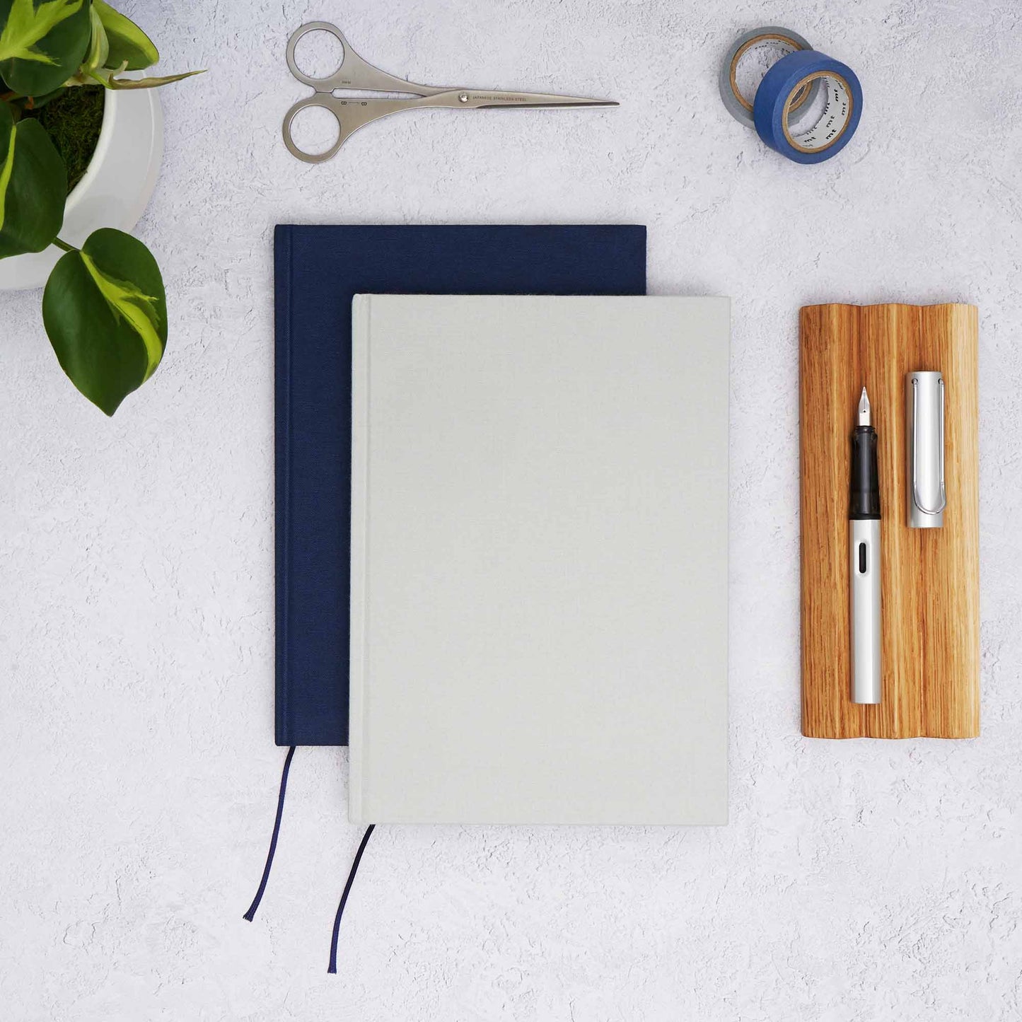 The Paper Mind Passepied Cream Hardcover Notebooks in grey and blue with an oak wood pen tray, Lamy AL Star fountain Pen, MT washi tape, Before Breakfast London scissors and plant.
